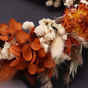 May include: A floral crown composed of dried and preserved botanical elements. The arrangement features vibrant orange hydrangea petals, clusters of small white baby's breath, fluffy beige bunny tail grass, and deep rust-colored dried flowers.