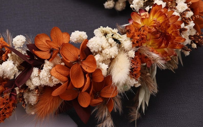 May include: A floral crown composed of dried and preserved botanical elements. The arrangement features vibrant orange hydrangea petals, clusters of small white baby's breath, fluffy beige bunny tail grass, and deep rust-colored dried flowers.