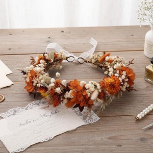 May include: A circular floral crown composed of dried orange and rust-colored flowers, white baby's breath, and tan grasses. The crown features a white lace ribbon tie at the back, creating a delicate, circular arrangement of preserved botanical elements.