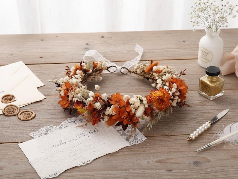 May include: A circular floral crown composed of dried orange and rust-colored flowers, white baby's breath, and tan grasses. The crown features a white lace ribbon tie at the back, creating a delicate, circular arrangement of preserved botanical elements.