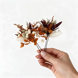 May include: Two hair pins featuring arrangements of dried flowers and foliage in shades of burnt orange, deep burgundy, cream, and sage green. Each pin has a thin black metal base with a U-shaped design to secure the floral clusters in place.