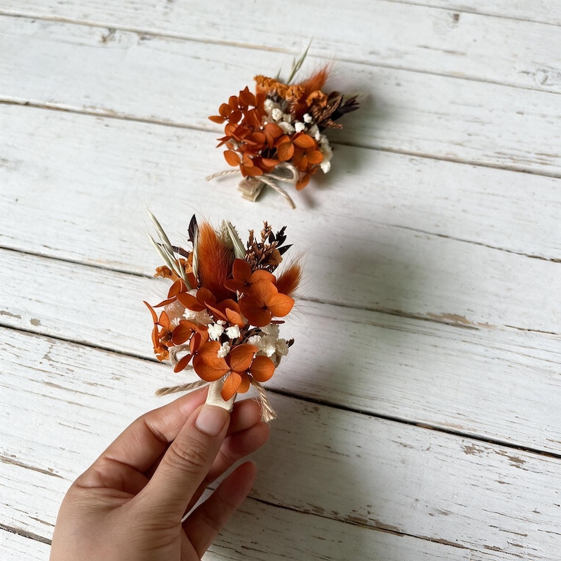May include: Two small boutonnieres featuring dried orange hydrangea petals, white baby's breath, and brown bunny tail grass. Each floral arrangement is secured with a light-colored wooden clip and tied with a thin piece of natural twine for attachment.