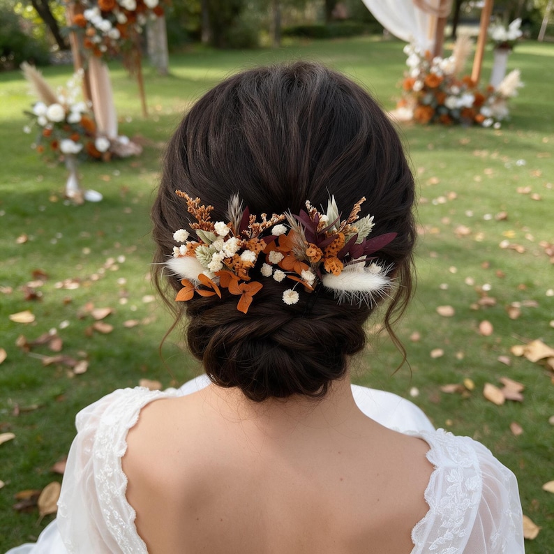May include: A hair accessory featuring a cluster of dried flowers in shades of burnt orange, deep burgundy, and cream. The arrangement includes small white baby's breath, fluffy pampas grass, and preserved foliage, secured into a dark brown updo.