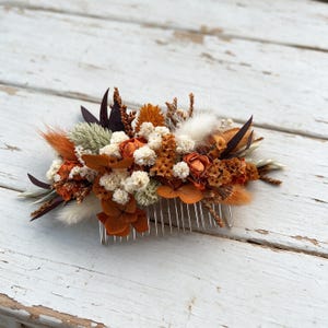 May include: A hair comb featuring an arrangement of dried and preserved botanical elements in shades of burnt orange, cream, and dark brown. The floral cluster includes small white flowers, orange petals, fluffy grass plumes, and dark pointed leaves.