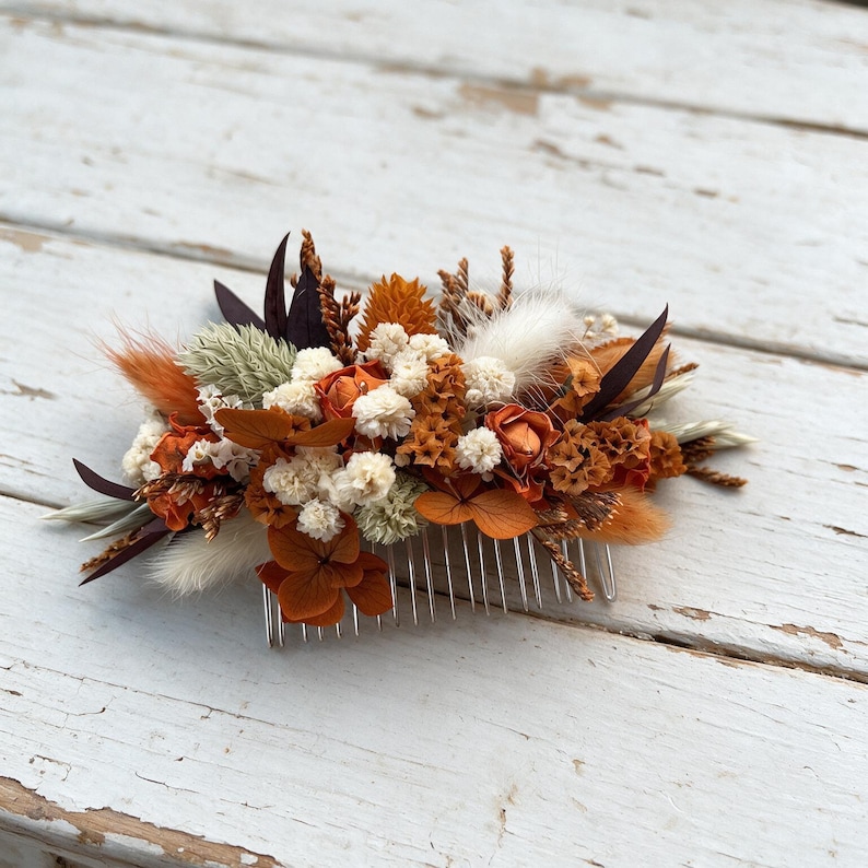 May include: A hair comb featuring an arrangement of dried and preserved botanical elements in shades of burnt orange, cream, and dark brown. The floral cluster includes small white flowers, orange petals, fluffy grass plumes, and dark pointed leaves.