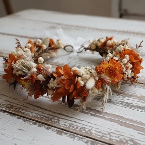 May include: A floral crown featuring a mix of dried and preserved flowers in shades of burnt orange, cream, and beige. The arrangement includes small white blossoms, orange petals, and feathery grasses, secured with a thin white ribbon at the back.