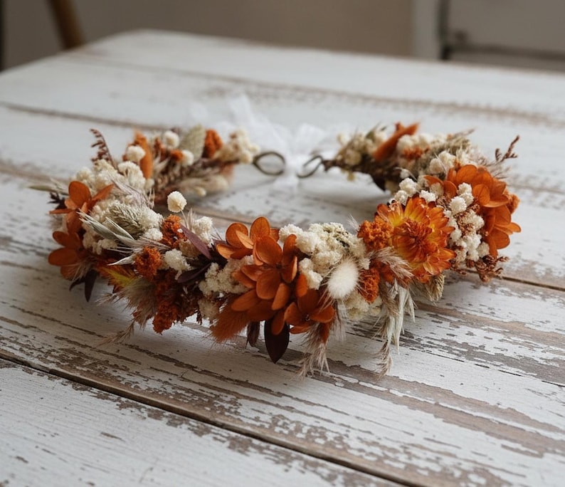 May include: A floral crown featuring a mix of dried and preserved flowers in shades of burnt orange, cream, and beige. The arrangement includes small white blossoms, orange petals, and feathery grasses, secured with a thin white ribbon at the back.