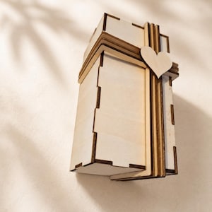 Könnte beinhalten: Eine rechteckige Schachtel aus hellem Holz mit ineinandergreifenden Verbindungen und einem dekorativen hölzernen Banddesign, das um die Außenseite gewickelt ist. Ein kleines, flaches hölzernes Herz ist mittig am Band befestigt.
