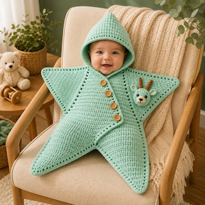 Op de afbeelding: Een licht mintgroene gehaakte babyslaapzak in de vorm van een vijfpuntige ster met een capuchon. Het kledingstuk is voorzien van vier ronde houten knopen langs de diagonale opening aan de voorzijde en een kleine gehaakte hertenkop applicatie.