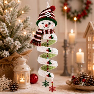 May include: A hanging crochet snowman ornament featuring a white spiral body, a striped red, green, and white hat with a pom-pom, and a matching scarf. The spiral is decorated with small green holly leaves, red berries, and colourful beads, ending with two snowflake charms.