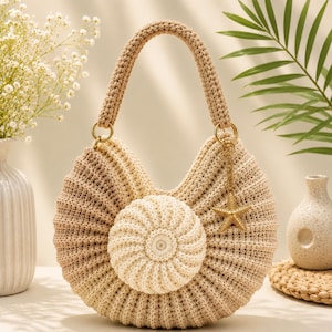 Peut inclure: Un sac à main en crochet en forme de croissant dans des tons beige et brun clair, doté d'une pièce centrale circulaire texturée, d'une anse tressée épaisse et d'une breloque étoile de mer en métal doré fixée à un anneau sur le côté.