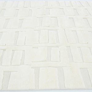 May include: A large, rectangular area rug in a solid off-white colour. The surface features a repeating geometric pattern of raised, textured rectangular blocks arranged in a grid-like formation, creating a subtle depth and tactile contrast.