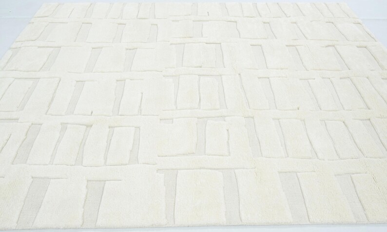 May include: A large, rectangular area rug in a solid off-white colour. The surface features a repeating geometric pattern of raised, textured rectangular blocks arranged in a grid-like formation, creating a subtle depth and tactile contrast.