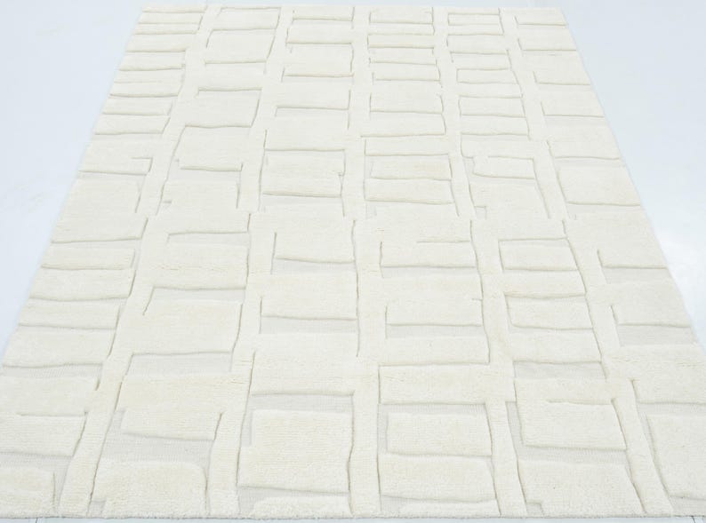 May include: A rectangular area rug in a solid off-white colour featuring a raised, textured geometric pattern of repeating rectangular blocks arranged in a grid-like formation across the entire surface of the floor covering.