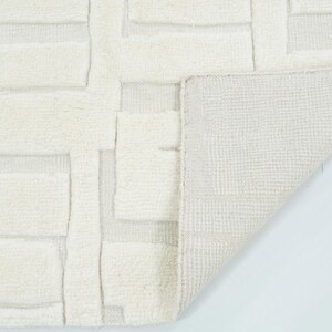 May include: An off-white area rug featuring a raised, geometric block pattern with a high-pile texture. The corner of the rug is folded over to reveal a flat, woven backing with a grid-like structure, contrasting with the plush, tufted surface.