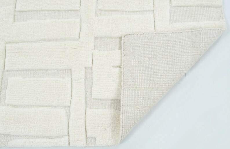 May include: An off-white area rug featuring a raised, geometric block pattern with a high-pile texture. The corner of the rug is folded over to reveal a flat, woven backing with a grid-like structure, contrasting with the plush, tufted surface.