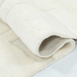 May include: A plush, off-white area rug featuring a textured, geometric block pattern. The rug is partially folded over, revealing a soft, thick pile on the top surface and a woven, light-coloured backing material on the underside of the fold.