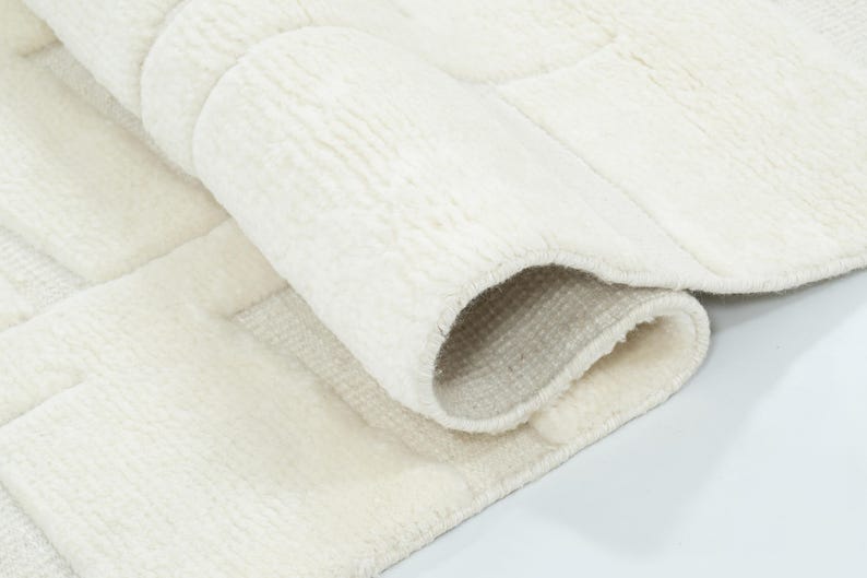 May include: A plush, off-white area rug featuring a textured, geometric block pattern. The rug is partially folded over, revealing a soft, thick pile on the top surface and a woven, light-coloured backing material on the underside of the fold.