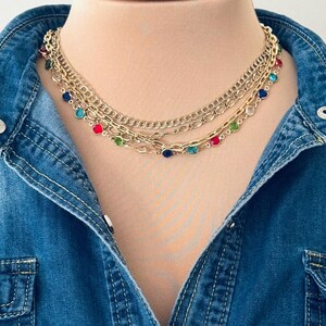 May include: A multi-strand gold-tone necklace featuring two plain metal chains and one chain accented with small, round, multi-colored gemstones in red, blue, green, and clear, worn over a blue denim shirt with silver-tone metal buttons.