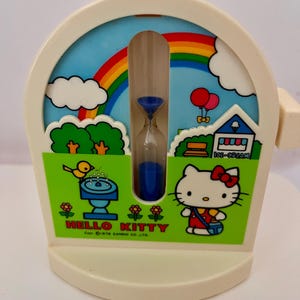 May include: A cream-coloured plastic timer featuring a central hourglass with blue sand. The graphic depicts Hello Kitty, a rainbow, clouds, a fountain, and an ice cream shop. The text reads HELLO KITTY and Cop. 1976 SANRIO CO. LTD.