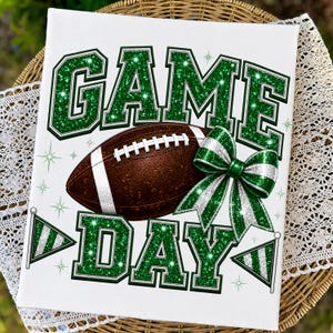 May include: A white rectangular sign featuring the text GAME DAY in large green glittery block letters. A brown football with white laces sits in the center, accompanied by a green and white glittery bow and two small pennant flags.