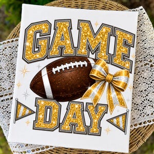 May include: A white rectangular sign featuring the text GAME DAY in large gold glitter block letters. A brown football with white laces sits in the center, accompanied by a gold and silver glitter bow and two small pennant flags on the sides.