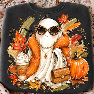 May include: A dark grey sweatshirt featuring a graphic of a white ghost wearing leopard print sunglasses, a gold chain necklace, and an orange faux fur stole. The ghost holds a quilted brown handbag next to a pumpkin, a coffee cup, and autumn leaves.