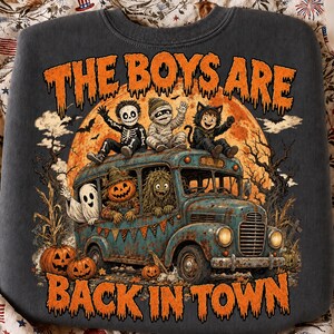 Puede incluir: Una sudadera gris carb&oacute;n con un gr&aacute;fico de un autob&uacute;s antiguo lleno de ni&ntilde;os disfrazados, incluyendo un esqueleto, momia, gato, fantasma, calabaza y monstruo. El texto THE BOYS ARE BACK IN TOWN aparece frente a una gran luna naranja.