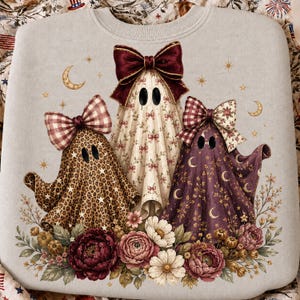 K&ouml;nnte beinhalten: Ein hellgraues Sweatshirt mit einer Grafik von drei Geistern, die mit gro&szlig;en Schleifen verziert sind. Der linke Geist hat ein Leopardenmuster, der mittlere ein Blumenmuster mit einer dunkelroten Schleife und der rechte ein violettes Mondmuster.