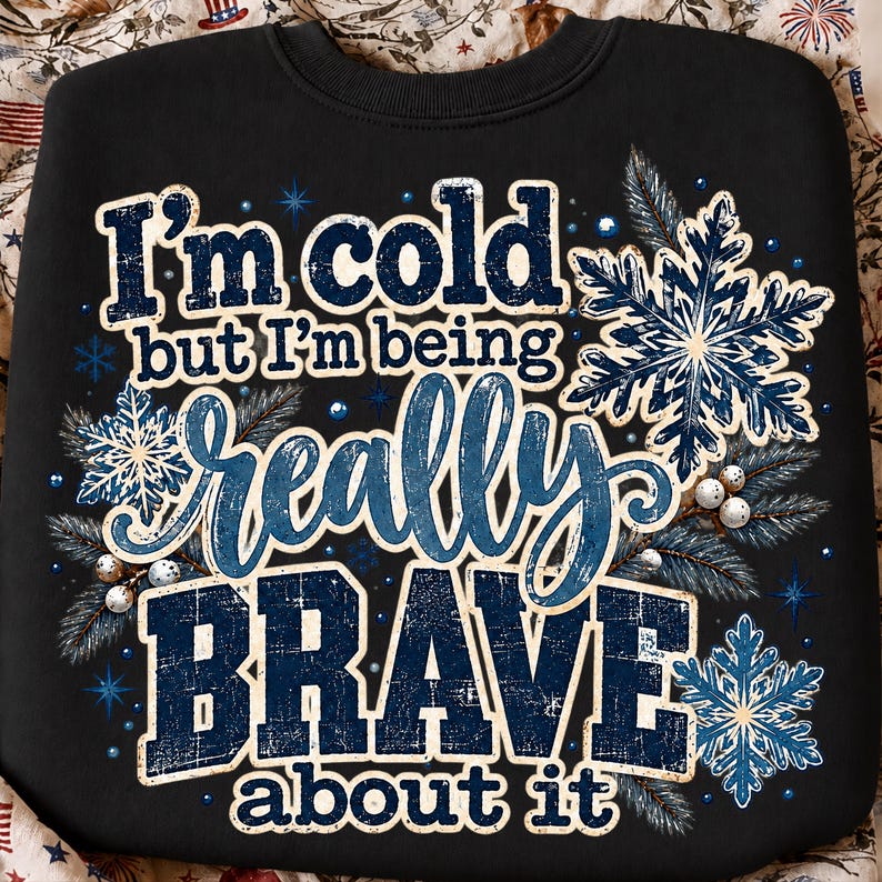 Może przedstawiać: Czarna bluza z napisem "I'm cold but I'm being really BRAVE about it" wykonanym niebiesko-białymi, postarzanymi literami, otoczona ilustracjami niebieskich płatk&oacute;w śniegu, gałązek sosny i małych białych jag&oacute;d.