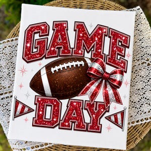 May include: A rectangular white sign featuring the text GAME DAY in large red glittery block letters. A brown football with white laces is centered between the words. A red and white striped bow and two small triangular pennants are included.