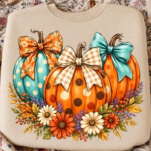 May include: A beige crewneck sweatshirt featuring a graphic print of three pumpkins. The left pumpkin is teal with white polka dots and an orange floral bow. The centre pumpkin is orange with brown polka dots and a beige gingham bow. The right pumpkin is orange with orange polka dots and a teal bow. The base is decorated with white and orange daisies, purple sprigs, and autumn leaves.