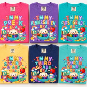 May include: Six Comfort Colors t-shirts in pink, purple, teal, yellow, navy, and light blue. Each features a graphic with the text In My Era, a smiling character in a crayon box, alphabet blocks, a rainbow, a red apple, and school supplies.