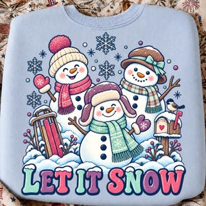 May include: A light blue sweatshirt featuring a graphic of three smiling snowmen wearing winter hats and scarves. The design includes snowflakes, a wooden sled, a mailbox with a heart, a small bird, and the text LET IT SNOW in colorful letters.