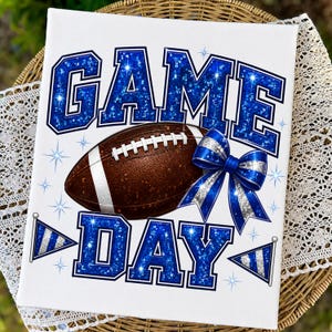 May include: A rectangular white canvas print featuring the text GAME DAY in large blue glitter letters. A brown football with white laces sits in the center, accented by a blue and silver striped bow and two small blue and silver pennant flags.