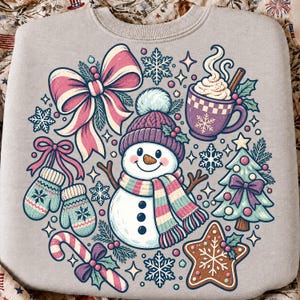 May include: A light gray crewneck sweatshirt featuring a central graphic of a snowman wearing a purple knit hat and a striped scarf, surrounded by a large pink bow, a mug of hot cocoa, a decorated Christmas tree, mittens, and a gingerbread star.