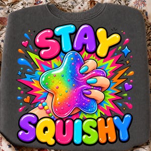 May include: A dark gray sweatshirt featuring a vibrant graphic of a hand squeezing a rainbow-colored, glittery star-shaped slime. The text STAY SQUISHY is printed in large, colorful, bubble-style letters above and below the central illustration.