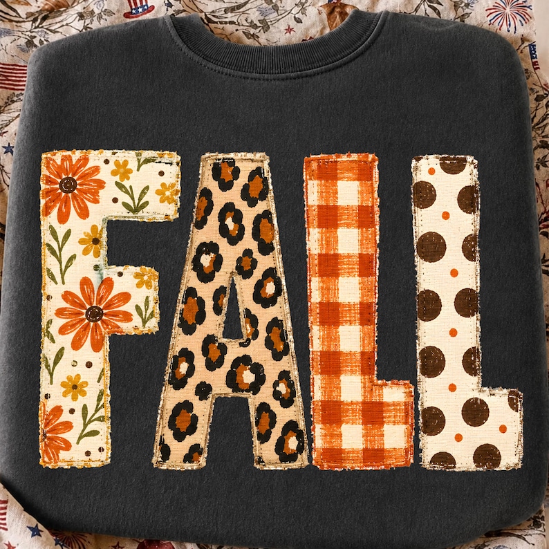 May include: A dark charcoal grey crewneck sweatshirt featuring the word FALL in large block letters. Each letter has a unique pattern: the F has orange flowers, the A has a leopard print, the L has orange gingham, and the L has brown polka dots.