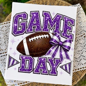 May include: A rectangular white sign featuring the text GAME DAY in large purple glitter letters. A brown football with white laces is centered between the words. A purple and silver striped bow and two small pennant flags are included.