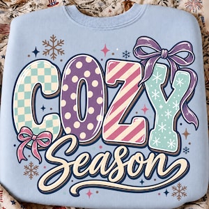 May include: A light blue sweatshirt featuring the text "COZY Season" in large, decorative letters. The word "COZY" has a checkered, polka dot, striped, and snowflake pattern, accented by two purple and pink bows. Small snowflakes and stars surround the text.