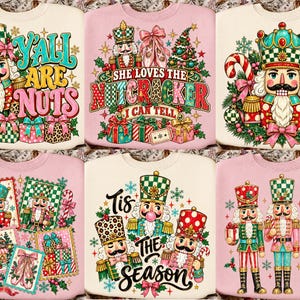 May include: A collection of six light-colored sweatshirts featuring colorful graphic prints of nutcrackers, Christmas presents, ballet slippers, and holiday greenery. The designs include text such as YALL ARE NUTS, SHE LOVES THE NUTCRACKER I CAN TELL, and TIS THE SEASON, rendered in various decorative fonts.