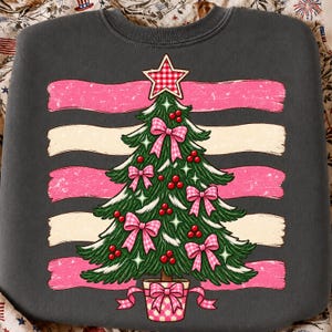 May include: A charcoal gray crewneck sweatshirt featuring a graphic of a green Christmas tree decorated with pink gingham bows, red berries, and a matching gingham star topper. The tree is set against horizontal pink and cream distressed brushstrokes.