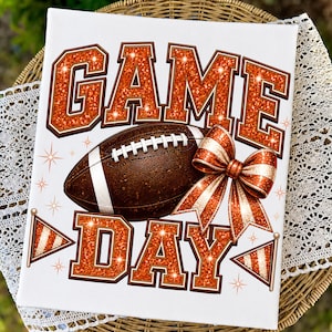 May include: A white rectangular sign featuring the text GAME DAY in large orange glitter block letters. A brown football with white laces is positioned between the words. An orange glitter bow and two small pennant flags decorate the design.