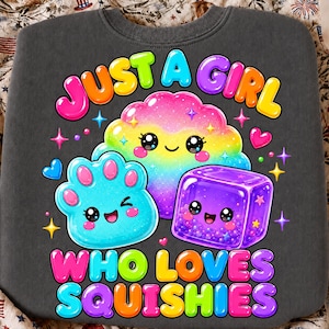 May include: A dark gray sweatshirt featuring a colorful graphic with the text "JUST A GIRL WHO LOVES SQUISHIES". The design includes a rainbow-colored cloud, a blue paw-shaped character, and a purple cube, all with smiling faces and sparkles.