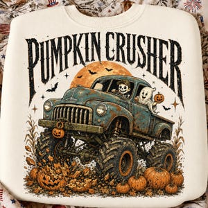 Puede incluir: Una sudadera color crema con el gr&aacute;fico de un cami&oacute;n monstruo azul con neum&aacute;ticos grandes pasando sobre calabazas. El texto PUMPKIN CRUSHER est&aacute; impreso arriba en una fuente oscura y desgastada. Un esqueleto y un fantasma van en el cami&oacute;n.