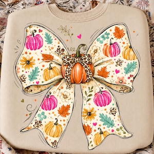 May include: A beige crewneck sweatshirt featuring a large central graphic of a bow tied around a pumpkin. The design includes orange, yellow, and pink pumpkins, autumn leaves, flowers, and leopard print accents on a light cream background.