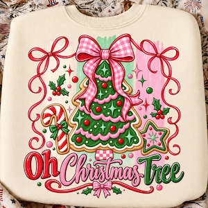 May include: A cream-colored sweatshirt featuring a graphic of a green Christmas tree shaped like a cookie with pink icing, topped with a large pink gingham bow. The design includes a candy cane, a star cookie, holly, and the text Oh Christmas Tree.