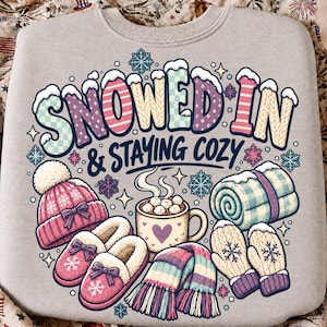 May include: A light gray sweatshirt featuring a colorful graphic with the text SNOWED IN & STAYING COZY. The design includes a pink knit hat, pink slippers, a mug of hot cocoa with marshmallows, a rolled blue blanket, and winter mittens.