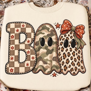 K&ouml;nnte beinhalten: Ein cremefarbenes Sweatshirt mit dem Wort BOO als Grafikdesign. Der Buchstabe B hat ein braun-wei&szlig;es Schachbrettmuster mit kleinen roten Blumen. Die beiden Os sind Geisterformen, eines mit gr&uuml;nem Tarnmuster und eines mit Leopardenmuster, wobei letzteres eine gro&szlig;e orangefarbene gepunktete Schleife tr&auml;gt.