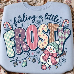 May include: A light blue crewneck sweatshirt featuring the text "feeling a little FROSTY" in dark blue script and large, multi-patterned block letters. A snowman wearing a teal beanie and pink scarf is positioned below the text.